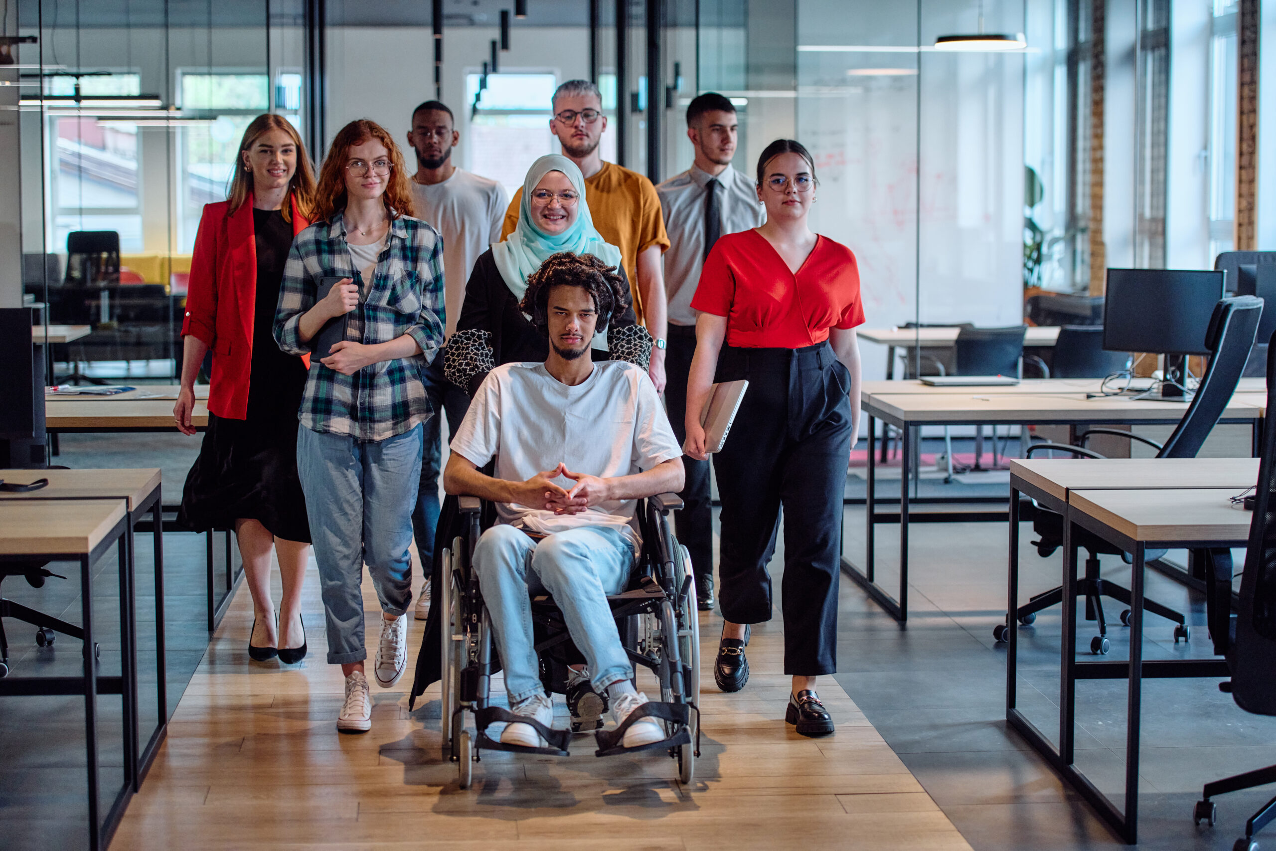 Un groupe diversifié de jeunes hommes d'affaires se rassemble dans le bureau vitré d'une startup moderne, mettant en vedette une personne en fauteuil roulant, un jeune homme afro-américain et une femme musulmane voilée.
